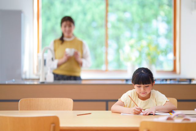 ダイニングテーブルで勉強する娘と、それをキッチンから見守る母親